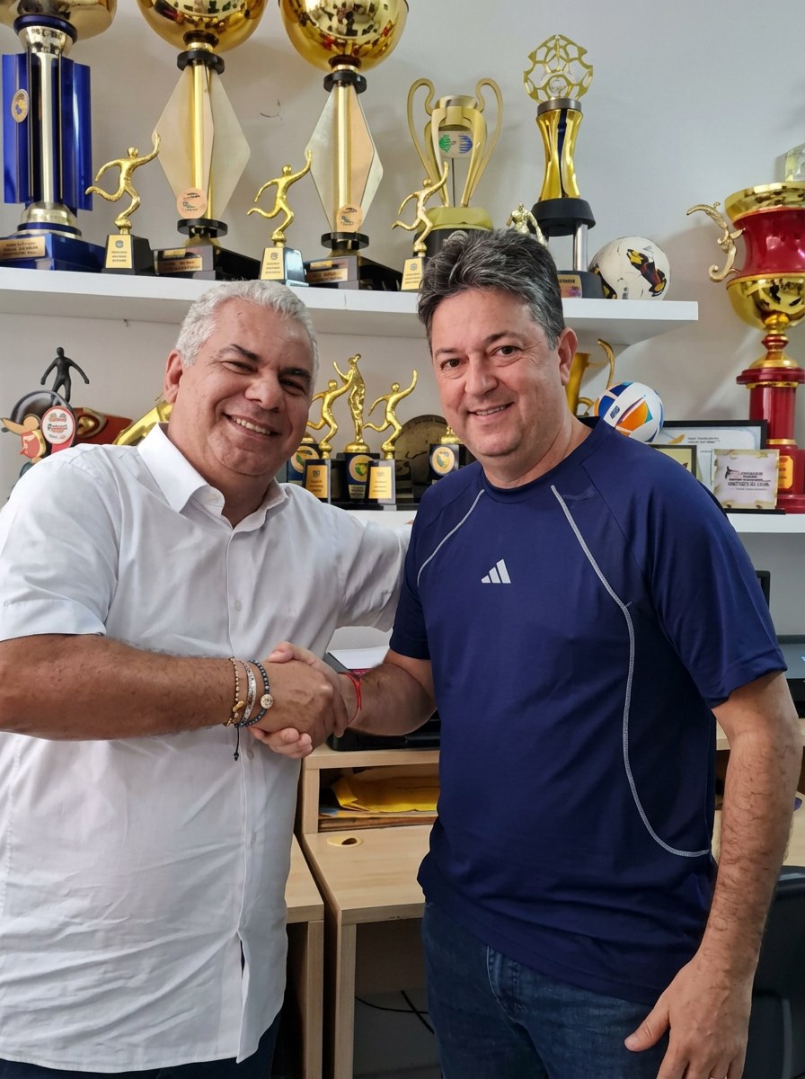 Adriano Pacheco com dirigente esportivo diante de uma estante de troféus